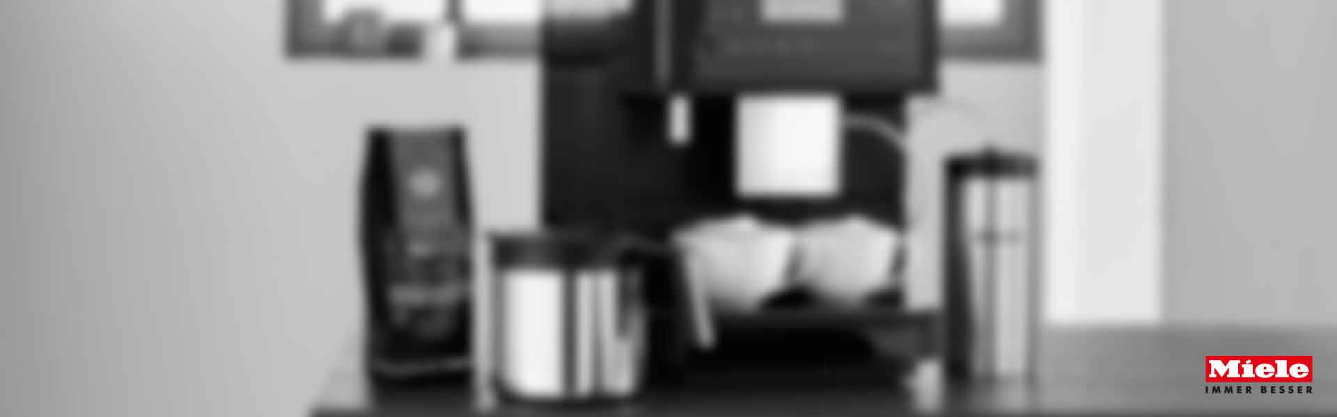 Eine Miele Kaffeemaschine mit vier Kaffeesorten, Kaffeebohnen und zwei Tassen Kaffee auf dunklem Hintergrund.